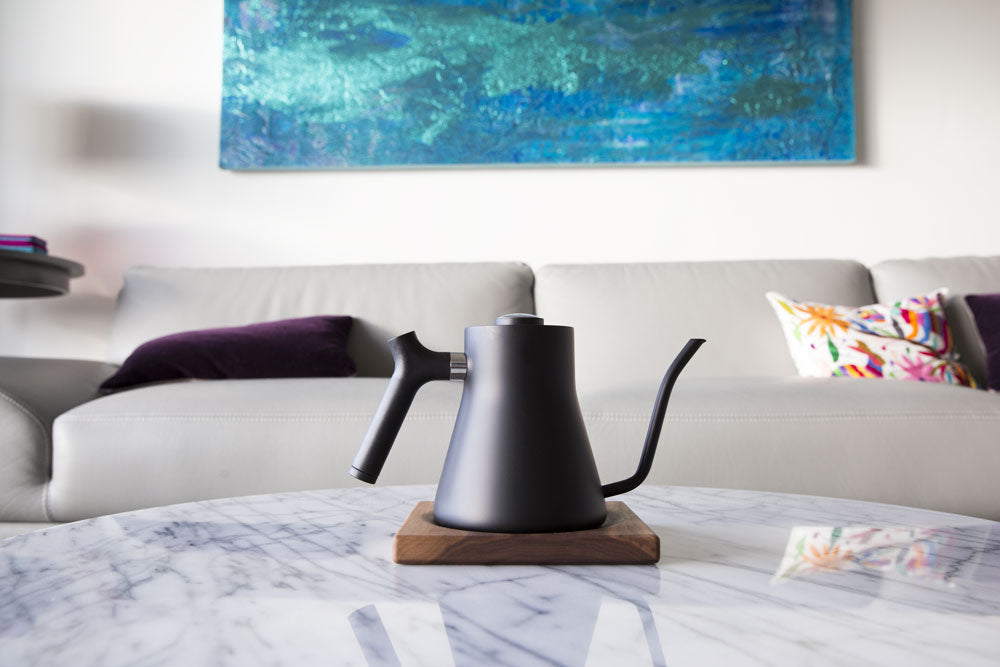  The Corvo EKG Pro electric kettle sits on a dining room coffee table, with
a couch and painting behind it.
