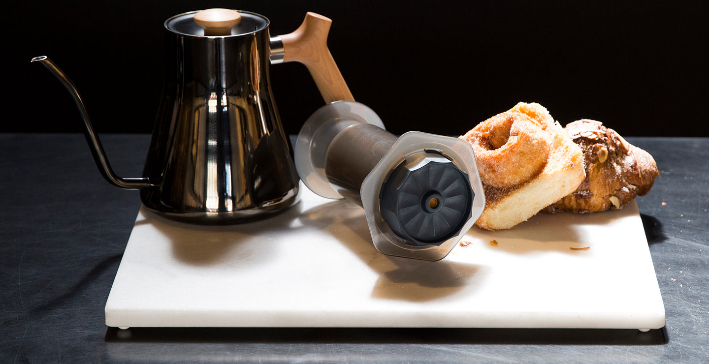 The Prismo attachment for AeroPress laying on a cutting board next to baked
goods and a Fellow kettle.
