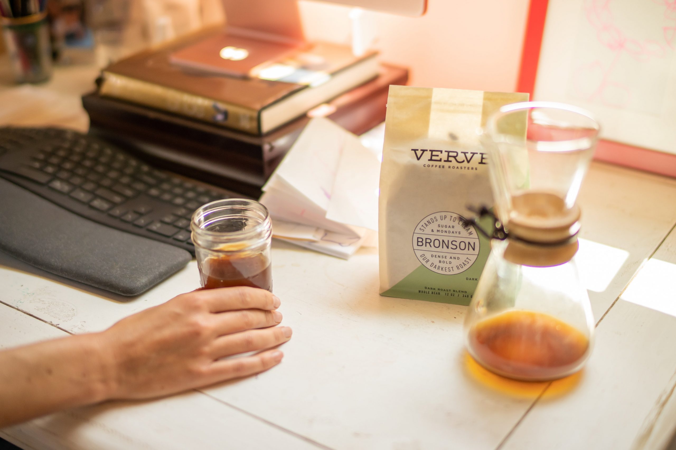 一袋 Verve Coffee 的 Bronson 法式烤糖蜜黑巧克力无花果味放在滴滤式咖啡机旁边的桌子上，一只手拿着一杯咖啡，还有一个键盘。