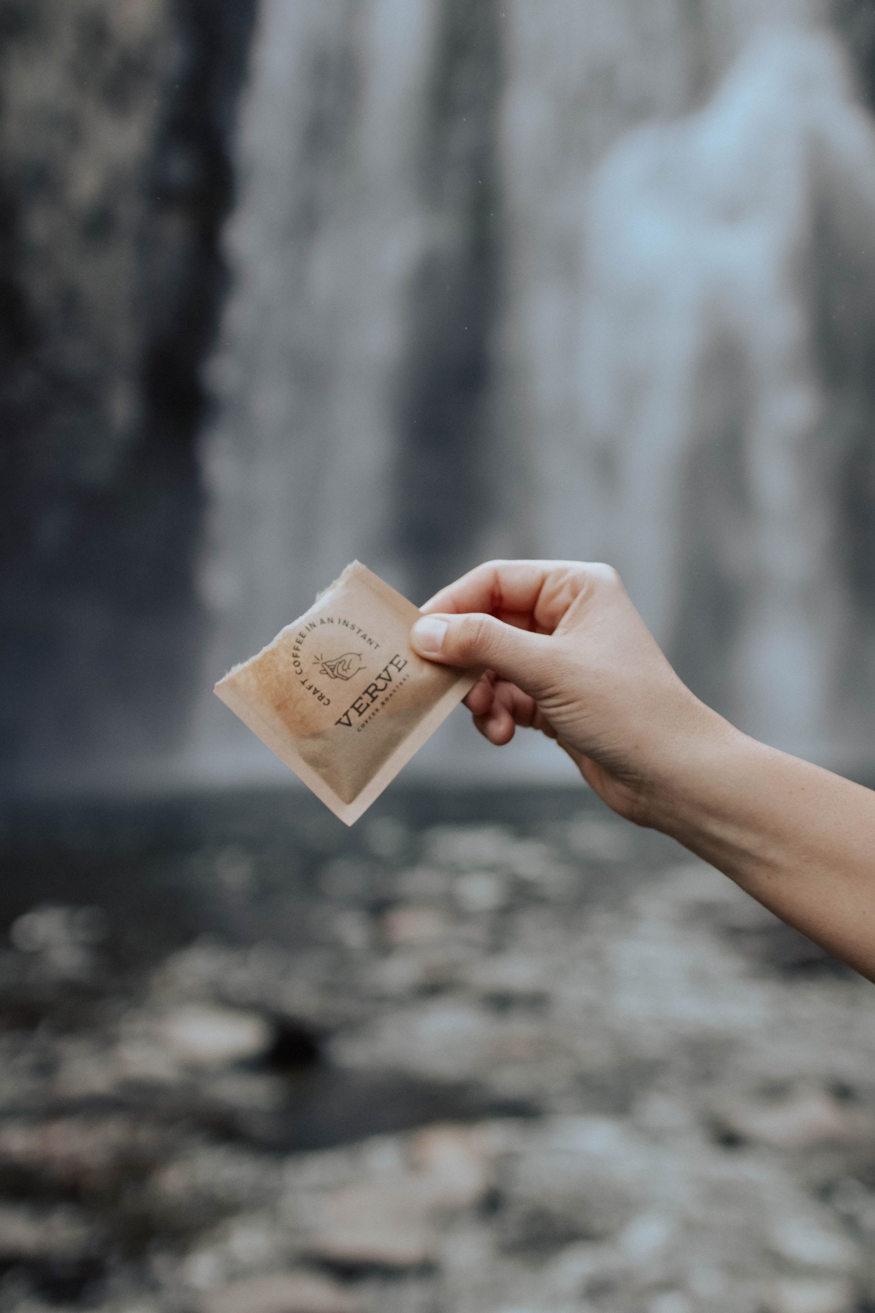 一只手拿着一袋打开的 Verve Coffee 手工速溶咖啡，站在湍急的瀑布前。